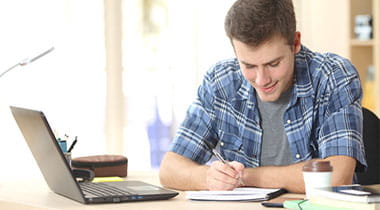 Young man sitting with laptop on desk, taking notes